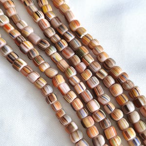 Può includere: Molteplici fili di piccole perline cilindriche disposte in linee parallele. Ogni perlina presenta un colore di base pesca chiaro o marrone chiaro con strisce verticali multicolori nelle tonalità del marrone, arancione, giallo, verde e blu.
