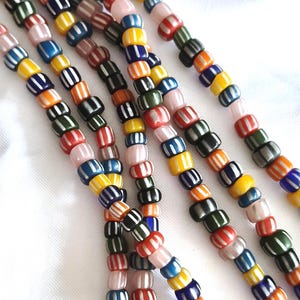 Op de afbeelding: Meerdere strengen cilindrische glaskralen met verticale witte strepen. De kralen zijn uitgevoerd in diverse kleuren, waaronder geel, blauw, oranje, groen, rood, zwart en lichtroze, wat zorgt voor een levendig en gestructureerd patroon.