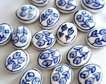 May include: A collection of oval-shaped ceramic beads, each featuring a white base with a hand-painted blue floral pattern depicting two tulips. The beads are arranged in a scattered formation, showcasing the consistent blue and white colour scheme.