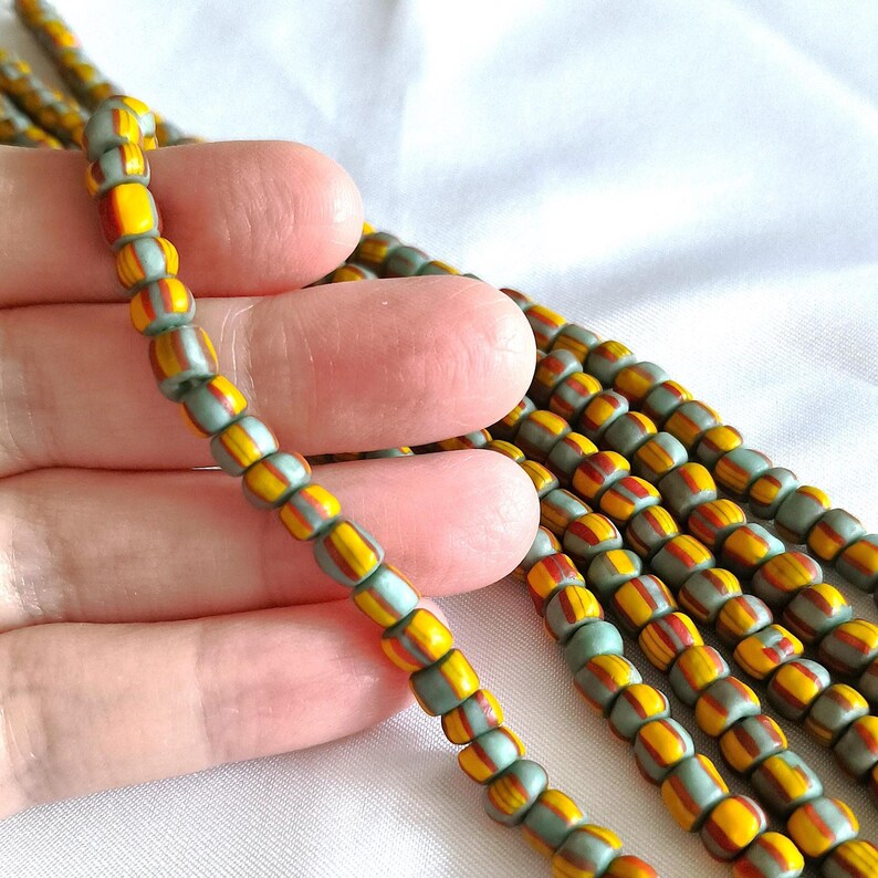 May include: A strand of small, cylindrical glass beads featuring a repeating pattern of muted sage green, vibrant yellow, and deep orange vertical stripes. The beads are strung together in multiple parallel rows, showcasing a consistent design.