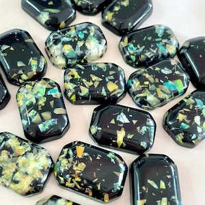 Può includere: Una collezione di cabochon in vetro nero e iridescente con un motivo maculato. I cabochon sono di varie forme e dimensioni.