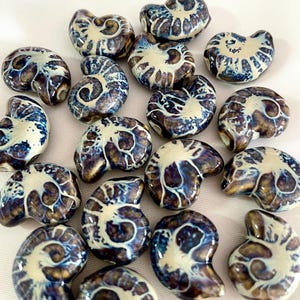 May include: A collection of ceramic ammonite beads. Each bead features a spiral design with a cream-coloured centre and a dark blue and brown exterior. The beads are arranged on a white surface.