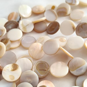 Puede incluir: Primer plano de una colección de cuentas redondas y planas de nácar en varios tonos de blanco, crema y marrón. Las cuentas están ensartadas, mostrando su brillo natural y sutiles variaciones de color. Ideal para la fabricación de joyas.