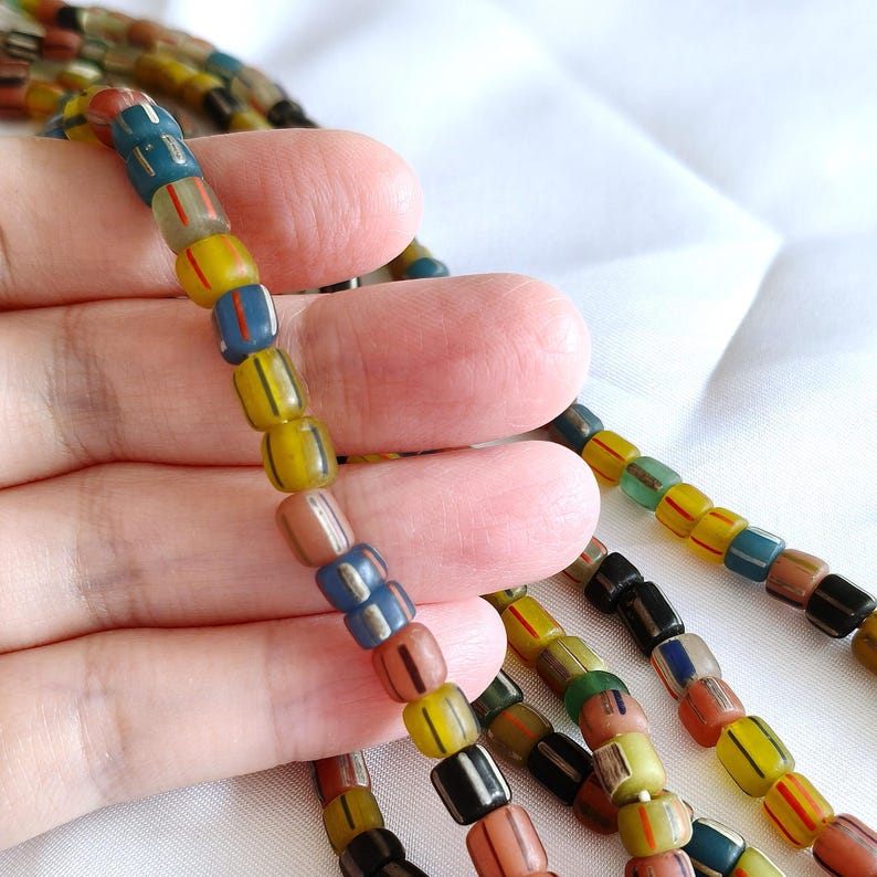 May include: A strand of cylindrical glass beads featuring a variety of colours including yellow, blue, brown, and black. Each bead is decorated with thin, vertical stripes in contrasting colours, arranged in a repeating pattern along the string.