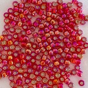 Puede incluir: Un montón grande de pequeñas cuentas de vidrio redondas, rojas y naranjas iridiscentes. Las cuentas tienen un agujero plateado en el centro.