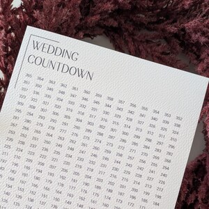 Puede incluir: Una tarjeta blanca de cuenta regresiva de boda con una corona floral burdeos. La tarjeta presenta una cuadrícula de números y el texto "WEDDING COUNTDOWN" y "one year until we say yes."