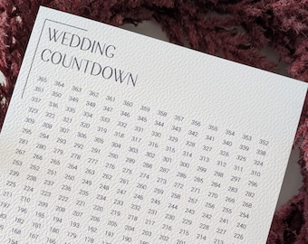 May include: A white wedding countdown card with a burgundy floral wreath. The card features a grid of numbers and the text "WEDDING COUNTDOWN" and "one year until we say yes."