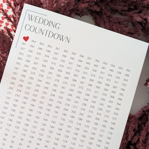 May include: A white wedding countdown calendar with a heart graphic, surrounded by a burgundy floral wreath. The calendar features a grid with numbers counting down from 364 to 2. The text "one year until we say yes" is at the bottom.