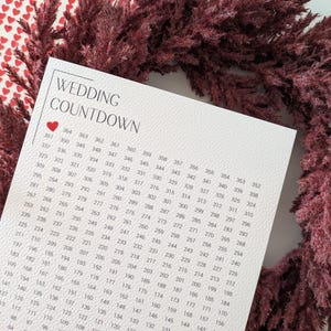 May include: A wedding countdown calendar with a heart graphic, surrounded by burgundy-colored dried flowers. The calendar features a grid of numbers counting down from 364 to 1, perfect for tracking the days until the wedding.
