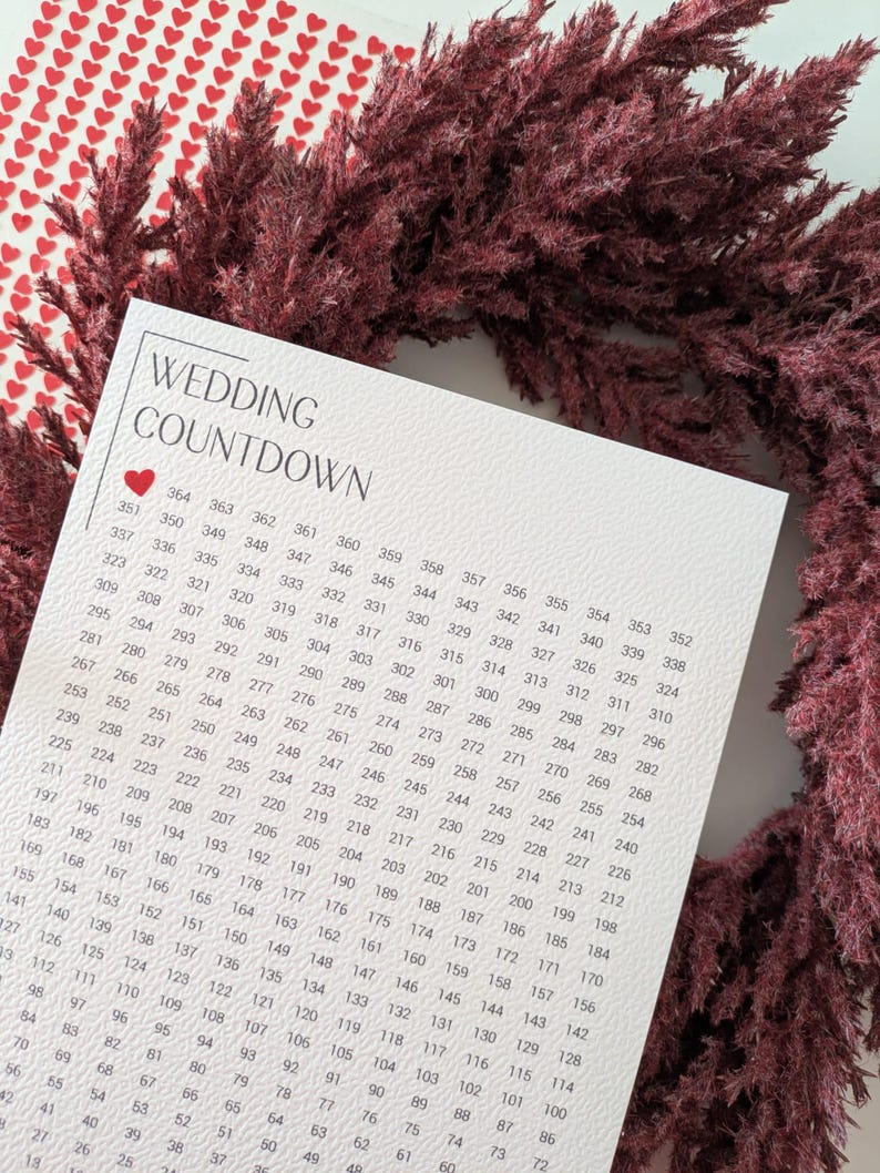 May include: A wedding countdown calendar with a heart graphic, surrounded by burgundy-colored dried flowers. The calendar features a grid of numbers counting down from 364 to 1, perfect for tracking the days until the wedding.