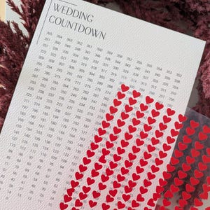 May include: A wedding countdown calendar with a white background and a grid of numbers. Red heart stickers are placed on the calendar. The text "WEDDING COUNTDOWN" is at the top.