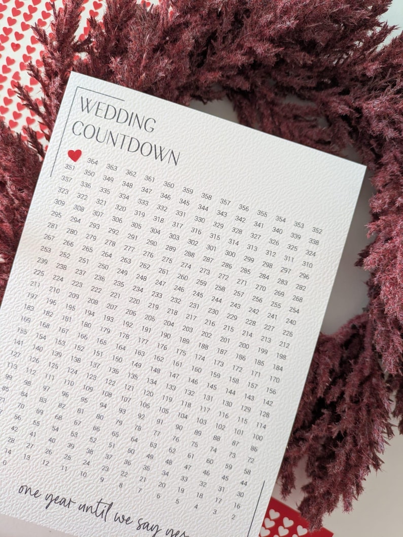 May include: A white wedding countdown calendar with a heart graphic, surrounded by a burgundy floral wreath. The calendar features a grid with numbers counting down from 364 to 2. The text "one year until we say yes" is at the bottom.