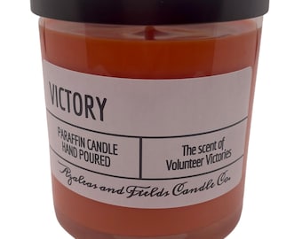 May include: A glass jar candle with a black lid. The candle is orange and has a white label that reads "VICTORY" and "PARAFFIN CANDLE HAND POURED". The label also says "The scent of Volunteer Victories" and "Azaleas and Fields Candle Co."