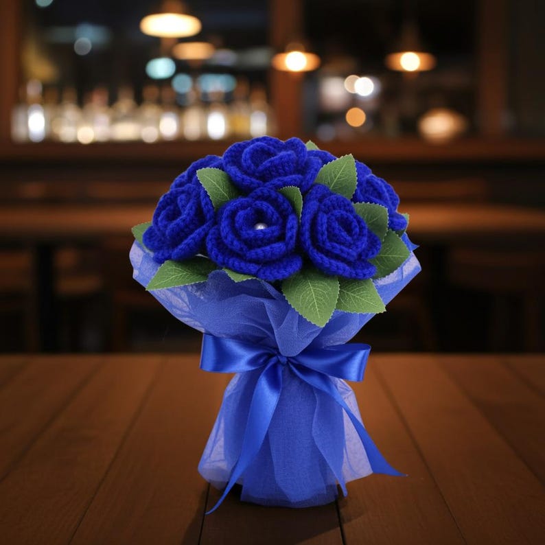 Puede incluir: Un ramo de rosas azules de ganchillo con hojas verdes, envuelto en papel de seda azul y atado con una cinta de raso azul. Una perla est&aacute; en el centro de cada rosa. El ramo est&aacute; sobre una mesa de madera.
