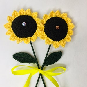 May include: Two handmade crochet sunflowers with yellow petals and dark centers, each adorned with a pearl-like bead. The stems are green, with green leaves and a yellow ribbon tied at the base. The sunflowers are a charming, handcrafted item.