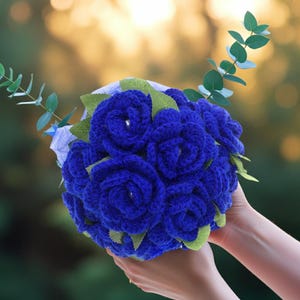 Puede incluir: Un ramo de rosas de ganchillo azul real hechas a mano con hojas verdes y ramitas de eucalipto. Las flores est&aacute;n dispuestas en forma redonda, sostenidas por manos sobre un fondo verde y dorado borroso.