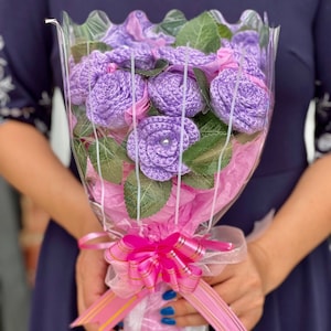 May include: A bouquet of crocheted purple roses, wrapped in clear cellophane and pink tissue paper. The bouquet is tied with a pink ribbon and bow. Green leaves are interspersed among the flowers, creating a vibrant and handmade floral arrangement.