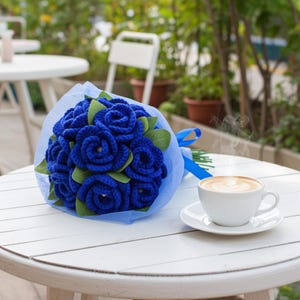 Puede incluir: Un ramo de rosas azules de ganchillo con hojas verdes, envuelto en papel de seda azul y atado con una cinta azul. Una taza de caf&eacute; y un platillo blancos est&aacute;n sobre una mesa blanca, con un fondo borroso al aire libre.