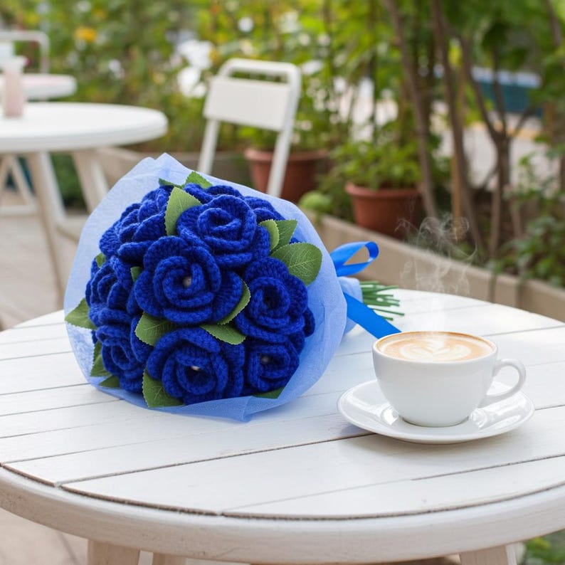 Puede incluir: Un ramo de rosas azules de ganchillo con hojas verdes, envuelto en papel de seda azul y atado con una cinta azul. Una taza de caf&eacute; y un platillo blancos est&aacute;n sobre una mesa blanca, con un fondo borroso al aire libre.