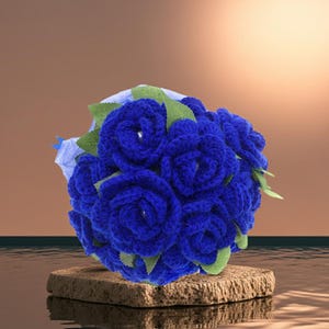 Puede incluir: Un ramo de rosas de ganchillo azul real hechas a mano con hojas verdes. Las flores est&aacute;n dispuestas en forma redonda y se presentan sobre una peque&ntilde;a piedra cuadrada texturizada de color marr&oacute;n claro.
