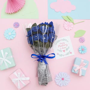 Puede incluir: Un ramo de rosas azules de fieltro envuelto en celof&aacute;n transparente y atado con una cinta azul. El ramo est&aacute; rodeado de flores de papel decorativas, cajas de regalo y una tarjeta que dice "Happy Mother's Day, a dear". El fondo es rosa.