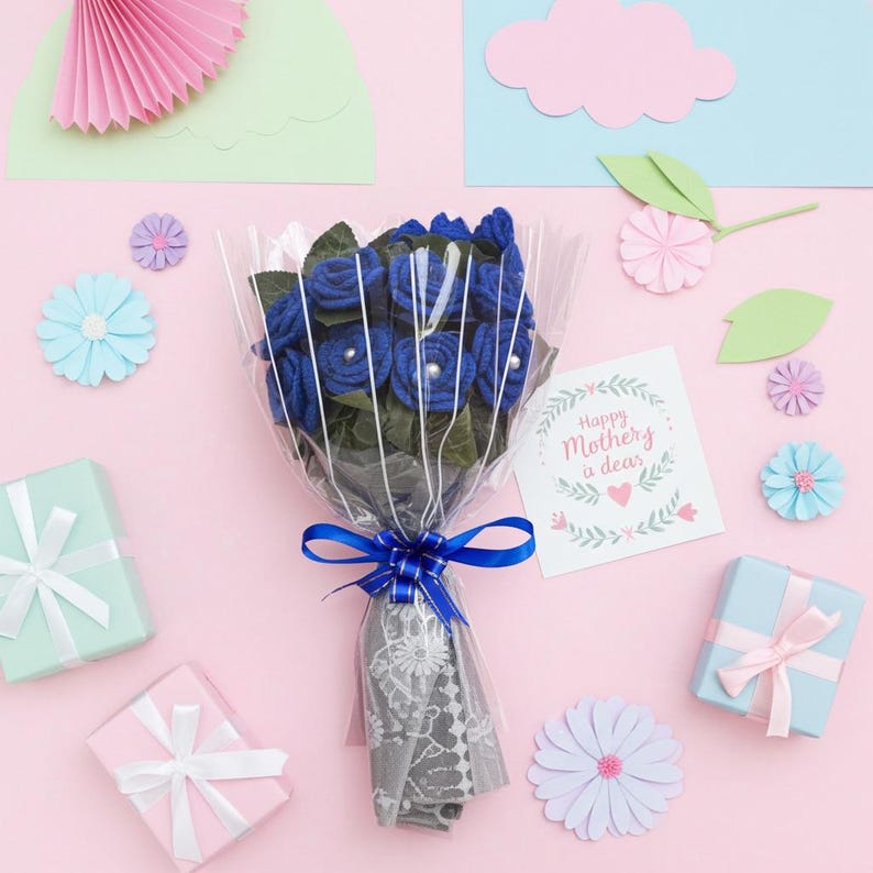 Puede incluir: Un ramo de rosas azules de fieltro envuelto en celof&aacute;n transparente y atado con una cinta azul. El ramo est&aacute; rodeado de flores de papel decorativas, cajas de regalo y una tarjeta que dice "Happy Mother's Day, a dear". El fondo es rosa.
