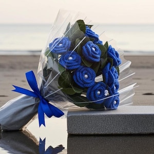 Puede incluir: Un ramo de rosas azules de ganchillo con centros de perlas, envuelto en celof&aacute;n transparente y atado con una cinta azul. El ramo se presenta sobre un bloque rectangular gris, con una playa y el oc&eacute;ano al fondo.