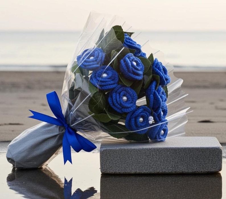 Puede incluir: Un ramo de rosas azules de ganchillo con centros de perlas, envuelto en celof&aacute;n transparente y atado con una cinta azul. El ramo se presenta sobre un bloque rectangular gris, con una playa y el oc&eacute;ano al fondo.