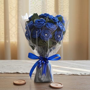 Puede incluir: Un ramo de rosas azules envuelto en celof&aacute;n transparente, atado con una cinta azul. Las rosas tienen detalles de perlas y hojas verdes. El ramo est&aacute; sobre una mesa de madera con dos botones de madera.
