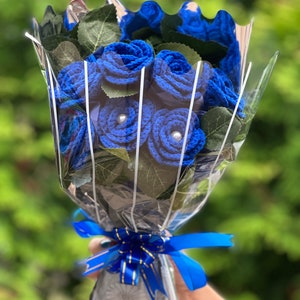 Puede incluir: Un ramo de rosas azules de ganchillo con centros de perlas, envuelto en celof&aacute;n transparente y atado con una cinta azul. El ramo incluye hojas verdes y se encuentra sobre un fondo verde borroso.
