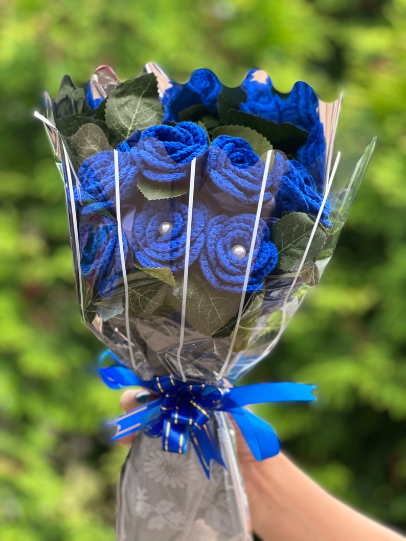 Puede incluir: Un ramo de rosas azules de ganchillo con centros de perlas, envuelto en celof&aacute;n transparente y atado con una cinta azul. El ramo incluye hojas verdes y se encuentra sobre un fondo verde borroso.