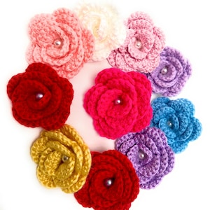 May include: A collection of handmade crochet flowers in various colors, including red, pink, yellow, and blue. Each flower features a pearl bead center. The flowers are arranged in a circular pattern against a white background.