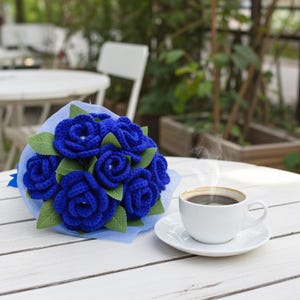 Puede incluir: Un ramo de rosas de ganchillo azul real hechas a mano con hojas verdes, presentado sobre una malla azul claro. Una taza y un platillo blancos con caf&eacute; humeante se encuentran sobre una mesa de madera blanca, creando una escena acogedora.