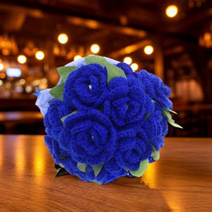 Puede incluir: Un ramo de rosas de ganchillo azul real hechas a mano con hojas verdes. Las flores est&aacute;n dispuestas en forma redonda, creando una apariencia completa y texturizada. El ramo est&aacute; envuelto en papel azul claro.
