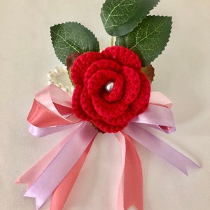 May include: A handmade red crochet rose with a pearl center, green leaves, and a pink and lavender ribbon bow. The floral arrangement is a decorative accent for weddings, parties, or special occasions.