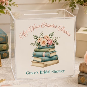 May include: A clear acrylic card box featuring the text "Her New Chapter Begins" and "Grace's Bridal Shower" with a central illustration of a stack of books topped with pink and peach roses and green leaves, designed for collecting event cards.