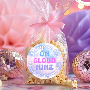 May include: A clear cellophane bag tied with a pink ribbon, filled with popcorn, and a round label that reads "Megan is on Cloud Nine". The label is 2 inches in diameter. Decorative disco balls and paper decorations are in the background.