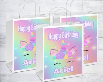 May include: Three white gift bags with unicorn designs and the text "Happy Birthday" and a name. The unicorn design features a rainbow background with a unicorn face, flowers, and glitter.