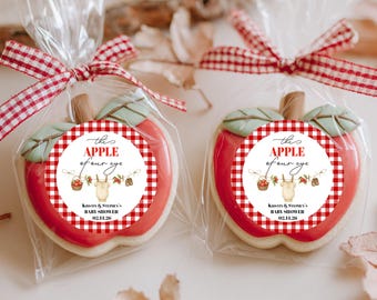 May include: Two apple-shaped cookies in clear cellophane bags, tied with red and white checkered ribbons. Each cookie has a red and white checkered design with the text "the APPLE of our eye" and "KRISTY & STEPHEN'S BABY SHOWER 02.11.26".