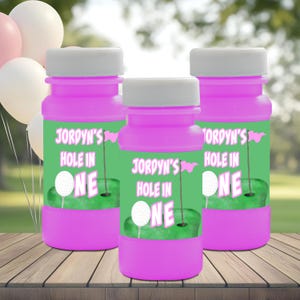 May include: Three pink bubble bottles with white caps and green labels. The labels read "Jordyn's Hole in One" with a golf ball and flag graphic. The bottles are on a wooden surface with balloons in the background.