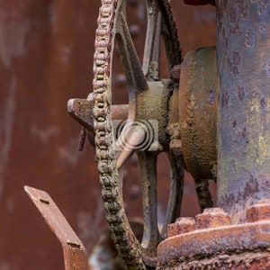 May include: A rusty metal gear with a chain attached to it. The gear is attached to a larger rusty metal pipe.