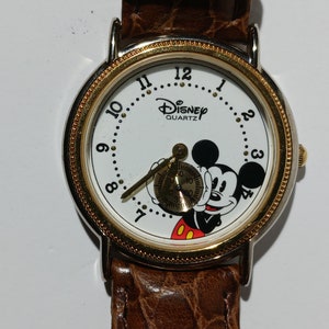 May include: A white Mickey Mouse watch with a brown leather band. The watch face features Mickey Mouse with the text "Disney Quartz" on the dial.