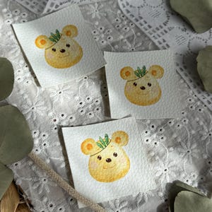 Könnte beinhalten: Drei quadratische Aufnäher mit einer Aquarellillustration eines gelben Bären mit einer grünen Blattkrone. Der Bär hat orange Ohren und eine kleine schwarze Nase und Augen. Die Aufnäher befinden sich auf einem weißen Spitzengrund mit grünen Blättern.