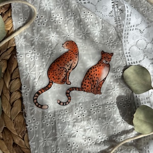 Könnte beinhalten: Zwei Aufkleber mit Geparden-Illustrationen. Die Geparden sind in sitzender Position mit orangefarbenem und schwarz geflecktem Fell dargestellt. Die Aufkleber befinden sich auf einem weißen Hintergrund mit floralem Muster und Spitzensaum.