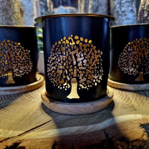 May include: Three black metal candle holders with a tree design cut out. The tree design is filled with a gold metallic finish. The candle holders are sitting on a wooden surface.
