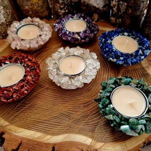 May include: Six tealight candle holders made of various semi-precious stones. The holders are arranged in a circle on a wooden surface. The stones include amethyst, lapis lazuli, clear quartz, red jasper, and green aventurine.