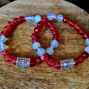 May include: Two bracelets with red and white beads. One bracelet has a silver charm with a symbol on it. The bracelets are on a wooden surface.