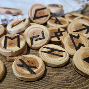 Könnte beinhalten: Eine Sammlung von Runensteinen aus Holz mit schwarz bemalten Symbolen. Die kreisförmigen Steine bestehen aus hellem Holz und zeigen die natürliche Maserung. Die Runen sind auf einer Holzoberfläche angeordnet und erzeugen eine rustikale und natürliche Ästhetik.