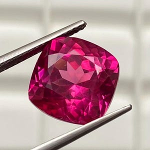 Puede incluir: Una gema vibrante, de talla cojín, en un tono magenta intenso. La superficie facetada refleja la luz, mostrando la claridad y el brillo de la gema. Sujeta con pinzas plateadas sobre un fondo blanco, resaltando su color y forma.