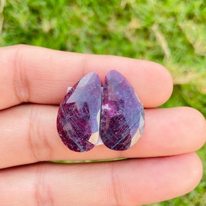 May include: Two pear-shaped ruby gemstones with a deep red and purple hue. The stones have a faceted cut and are set in a silver tone setting.